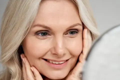 Uśmiechnięta 40-latka z blond włosami, patrząca w lustro. Delikatny makijaż na wesele podkreśla jej urodę.