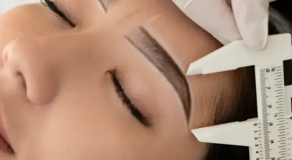 Precyzyjne pomiary przed laminacją brwi z farbką. Stylistka w białej rękawiczce mierzy łuk brwiowy.